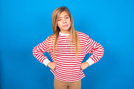 Photo for Funny and frustrated teen girl wearing striped shirt over blue background holding hands on waist and silly looking at awkward situation. - Royalty Free Image