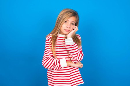 Photo for Very bored teen girl wearing striped shirt over blue background holding hand on cheek while support it with another crossed hand, looking tired and sick. - Royalty Free Image