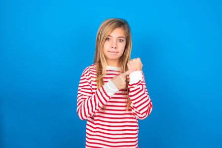 Téléchargez les photos : Adolescent fille portant rayé chemise sur fond bleu à la hâte pointant pour regarder le temps, impatience, bouleversé et en colère pour délai délai - en image libre de droit