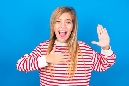 I swear, promise you not regret. Portrait of sincere teen girl wearing striped shirt over blue background raising one arm and hold hand on heart as give oath, telling truth, want you to believe.