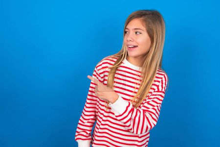 Téléchargez les photos : Joyeux souriant adolescent fille souriante portant chemise rayée sur fond bleu regardant et pointant de côté avec la main. Espace de copie et concept publicitaire. - en image libre de droit