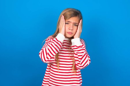 Photo for Teen girl wearing striped shirt over blue background holding head in hands with unhappy expression watching sad movie about animals and trying not to cry. - Royalty Free Image