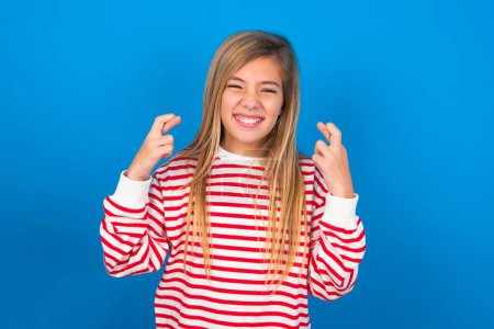 Téléchargez les photos : Adolescent fille portant chemise rayée sur fond bleu tenant les doigts croisés avec expression inquiète espérant patron n'a pas remarqué d'erreurs au travail. - en image libre de droit