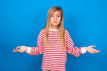 Foto de Adolescente chica usando rayas camisa sobre azul fondo looks incierto shrugs hombros. - Imagen libre de derechos