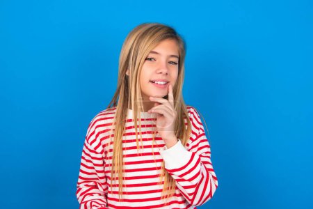 Photo for Carefree successful teen girl wearing striped shirt over blue background touching jawline gazing camera tilting head grinning white teeth delighted. Dental care concept. - Royalty Free Image