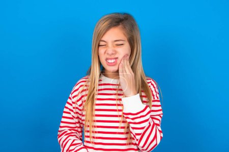 Photo for Toothache concept. teen girl wearing striped shirt over blue background feeling pain, holding his cheek with hand, suffering from bad toothache, looking at camera with painful expression - Royalty Free Image