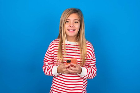 Téléchargez les photos : Souriant adolescent fille portant chemise rayée sur fond bleu amical et heureux tenant téléphone mobile prenant selfie dans le miroir. - en image libre de droit