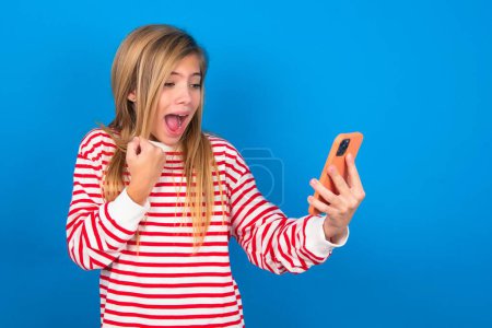 Téléchargez les photos : Heureuse adolescente joyeuse portant une chemise rayée sur fond bleu recevant de bonnes nouvelles par e-mail et célébrant le succès tout en se tenant debout et en regardant le téléphone mobile. - en image libre de droit