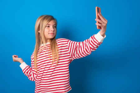 Foto de Retrato de una adolescente con camisa a rayas sobre fondo azul tomando una selfie para enviarla a amigos y seguidores o publicarla en sus redes sociales. - Imagen libre de derechos