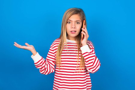 Photo for Teen girl wearing striped shirt over blue background talking on the phone stressed with hand on face, shocked with shame and surprise face, angry and frustrated. Fear and upset for mistake. - Royalty Free Image