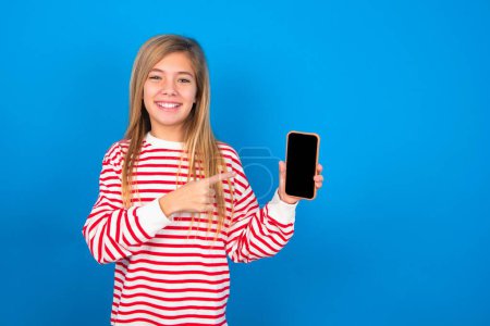 Foto de Chica adolescente sonriente con camisa a rayas sobre fondo azul que muestra y señala la pantalla del teléfono vacía. Concepto de publicidad y comunicación. - Imagen libre de derechos