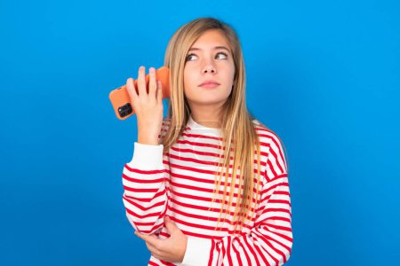 Photo for Smiling teen girl wearing striped shirt over blue background listening a voice message from her smartphone. Communication and technology concept. - Royalty Free Image