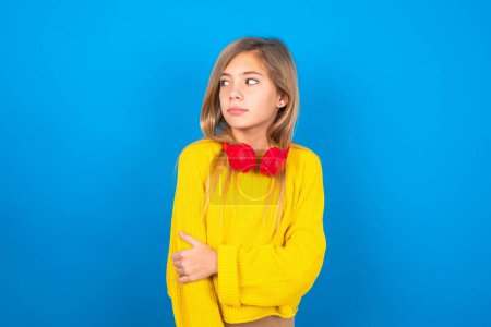 Téléchargez les photos : Plaisir caucasien adolescent fille portant pull jaune sur fond bleu studio garde les mains croisées sur la poitrine semble heureusement de côté - en image libre de droit