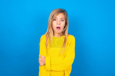 Photo for Shocked embarrassed caucasian teen girl wearing yellow sweater over blue studio background keeps mouth widely opened. Hears unbelievable novelty stares in stupor - Royalty Free Image