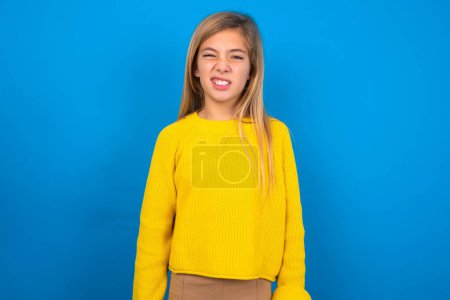 Foto de Retrato de chica adolescente caucásica insatisfecha usando suéter amarillo sobre fondo de estudio azul sonríe cara, bolsos labios y se ve con molestia en la cámara, descontento escuchando algo desagradable - Imagen libre de derechos