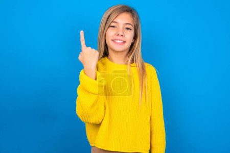 Téléchargez les photos : Souriant caucasien adolescent fille portant un pull jaune sur fond bleu studio montrant numéro un ou premier avec la main vers l'avant, compte à rebours - en image libre de droit