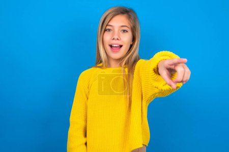 Téléchargez les photos : Excité positive caucasienne adolescent fille portant pull jaune sur fond bleu studio points index index directement à vous, voit quelque chose de très drôle. Wow, incroyable - en image libre de droit