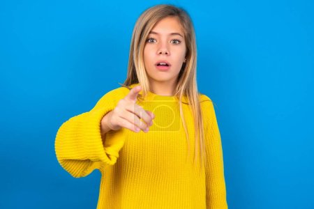 Téléchargez les photos : Choqué caucasien adolescent fille portant pull jaune sur bleu studio points de fond à vous avec une expression stupéfaite - en image libre de droit