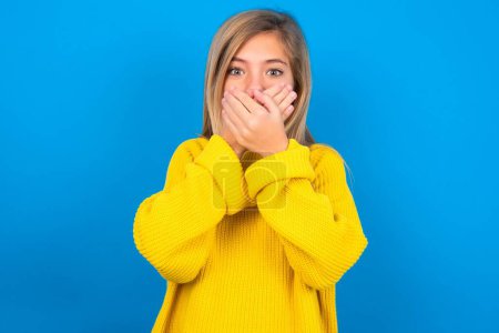 Photo for Stunned caucasian teen girl wearing yellow sweater over blue studio background covers mouth with both hands being afraid from something or after hearing stunning gossips. - Royalty Free Image