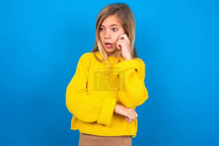 Foto de Sorprendida chica adolescente caucásica con suéter amarillo sobre fondo azul estudio se ve a un lado sorprendentemente con la boca abierta. - Imagen libre de derechos