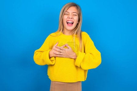 Foto de Hermosa chica adolescente caucásica con suéter amarillo sobre fondo de estudio azul expresa la felicidad, ríe agradablemente, mantiene las manos en el corazón - Imagen libre de derechos