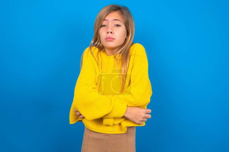 Téléchargez les photos : Belle fille adolescente caucasienne portant un pull jaune tremblant et gelant pour le froid hivernal avec une expression triste et choquante sur le visage sur fond bleu - en image libre de droit