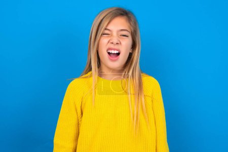 Foto de Chica adolescente caucásica con suéter amarillo sobre fondo de estudio azul bosteza con la boca abierta se levanta. Rutina diaria - Imagen libre de derechos
