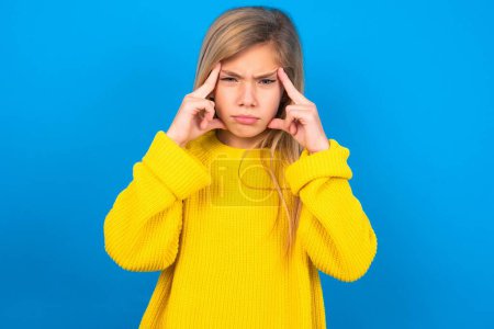 Photo for Serious concentrated caucasian teen girl wearing yellow sweater over blue wall keeps fingers on temples, tries to ease tension, gather with thoughts and remember important information for exam - Royalty Free Image