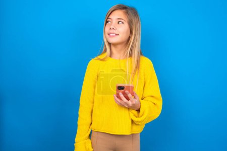 Foto de Rubia chica adolescente caucásica con suéter amarillo sobre fondo azul estudio sostiene el teléfono móvil utiliza Internet de alta velocidad y las redes sociales tiene comunicación en línea. Concepto de tecnologías modernas - Imagen libre de derechos