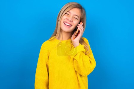 Foto de Divertido caucásico adolescente chica usando amarillo suéter sobre azul estudio ríe felizmente, tiene conversación telefónica, siendo divertido por amigo, cierra los ojos. - Imagen libre de derechos