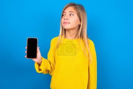Téléchargez les photos : Belle fille d'adolescence caucasienne portant un pull jaune sur fond bleu studio détient un nouveau téléphone mobile et semble mystérieux montre de côté affichage vierge de cellulaire moderne - en image libre de droit