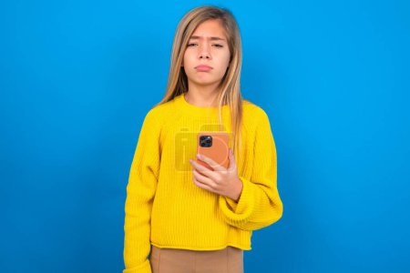 Foto de Muchacha adolescente caucásica trastornada que usa suéter amarillo sobre fondo azul del estudio utiliza la aplicación de software móvil y la información de navegación en Internet, sostiene el móvil moderno - Imagen libre de derechos