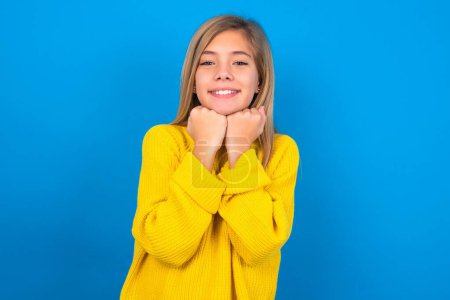 Foto de Satisfecho chica adolescente caucásica con suéter amarillo sobre fondo de estudio azul toca la barbilla con ambas manos, sonríe agradablemente, se regocija buen día con el amante - Imagen libre de derechos