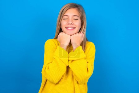 Foto de Alegre caucásico adolescente chica usando amarillo suéter sobre azul estudio fondo tiene tímido satisfecho expresión, sonrisas ampliamente, muestra dientes blancos, personas emociones - Imagen libre de derechos