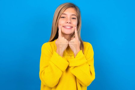 Foto de Feliz chica adolescente caucásica con suéter amarillo sobre fondo azul estudio con sonrisa dentada, mantiene los dedos índice cerca de la boca, dedos señalando y forzando sonrisa alegre - Imagen libre de derechos