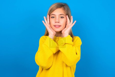 Photo for Happy caucasian teen girl wearing yellow sweater touches both cheeks gently, has tender smile, shows white teeth, gazes positively straightly at camera on blue background - Royalty Free Image