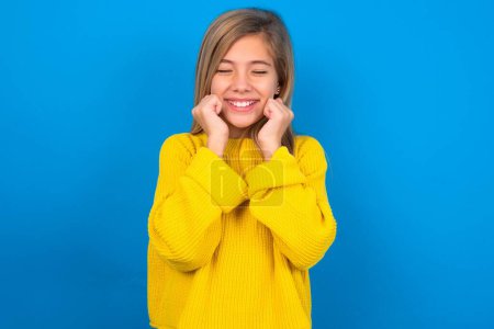Foto de Chica adolescente caucásica con suéter amarillo sobre fondo azul estudio sonríe alegremente, imagina algo agradable, espacio de copia. Concepto de emociones agradables. - Imagen libre de derechos