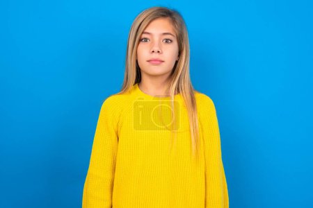 Téléchargez les photos : Une adolescente caucasienne stupéfaite portant un pull jaune sur un fond de studio bleu réagit aux nouvelles choquantes. Surpris fille retient souffle - en image libre de droit