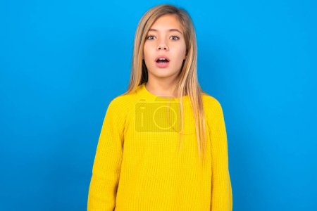 Shocked caucásico adolescente chica usando amarillo suéter sobre azul estudio fondo miradas con micrófonos ojos mantiene la boca abierta ha sorprendido a la expresión. Concepto de Omg 