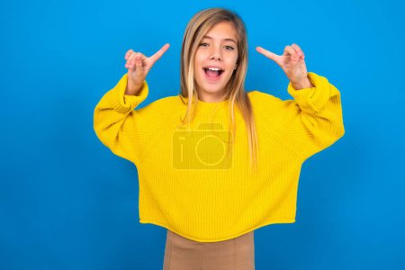 Téléchargez les photos : Joyeux caucasien adolescent fille portant pull jaune sur fond bleu studio démontrant coiffure - en image libre de droit