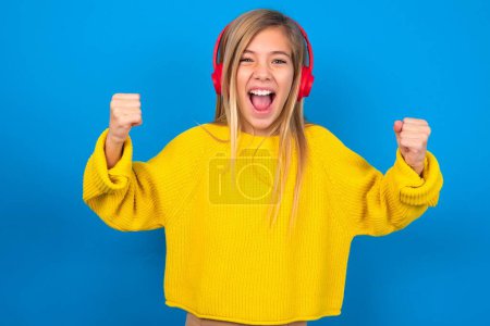 Photo for Emotional caucasian teen girl wearing yellow sweater over blue studio background  wears stereo headphones on ears makes yes gesture, listens favorite music - Royalty Free Image