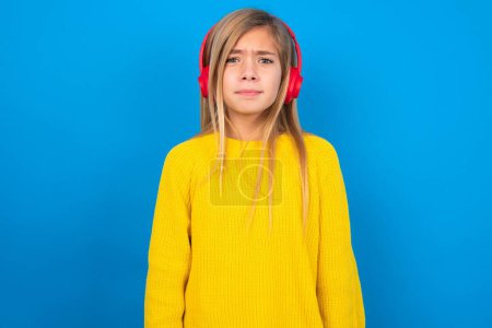 Téléchargez les photos : Mécontent caucasien adolescent fille porter jaune pull sur bleu studio arrière-plan semble perplexe à caméra être en colère porte casque stéréo écoute de la musique - en image libre de droit