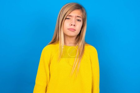 Foto de Descontenta chica adolescente caucásica usando suéter amarillo sobre fondo azul estudio frunce el ceño se siente infeliz tiene algunos problemas. Concepto de emociones y sentimientos negativos - Imagen libre de derechos