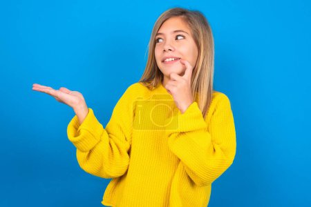 Téléchargez les photos : Positive caucasien adolescent fille porter jaune pull sur bleu studio arrière-plan annonce promo touches dents avec doigt. - en image libre de droit