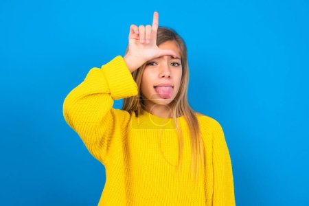 Photo for Funny caucasian teen girl wearing yellow sweater over blue studio background makes loser gesture mocking at someone sticks out tongue making grimace face. - Royalty Free Image