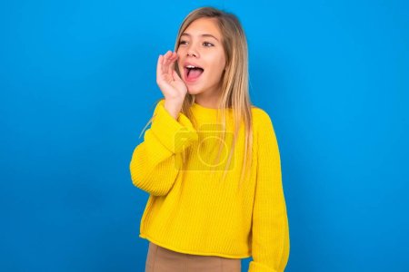 Foto de Hermosa chica adolescente caucásica usando suéter amarillo sosteniendo la mano cerca de su cara y gritando o llamando a alguien sobre fondo azul - Imagen libre de derechos