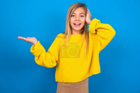 Téléchargez les photos : Surpris caucasien adolescent fille portant pull jaune sur fond bleu studio tient la main offrant proposition - en image libre de droit