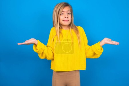 Téléchargez les photos : Caucasienne adolescent fille portant pull jaune sur fond bleu studio haussant les épaules, oups. - en image libre de droit