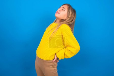 Photo for Beautiful caucasian teen girl wearing yellow sweater over blue studio background has back pain - Royalty Free Image