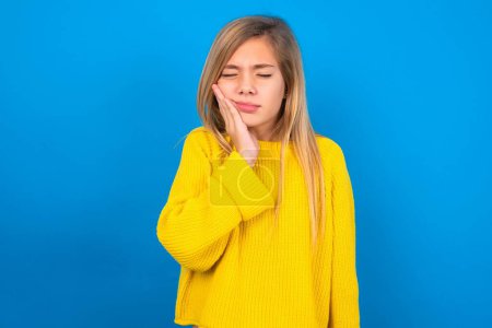 Photo for Blonde caucasian teen girl wearing yellow sweater over blue studio background with toothache - Royalty Free Image